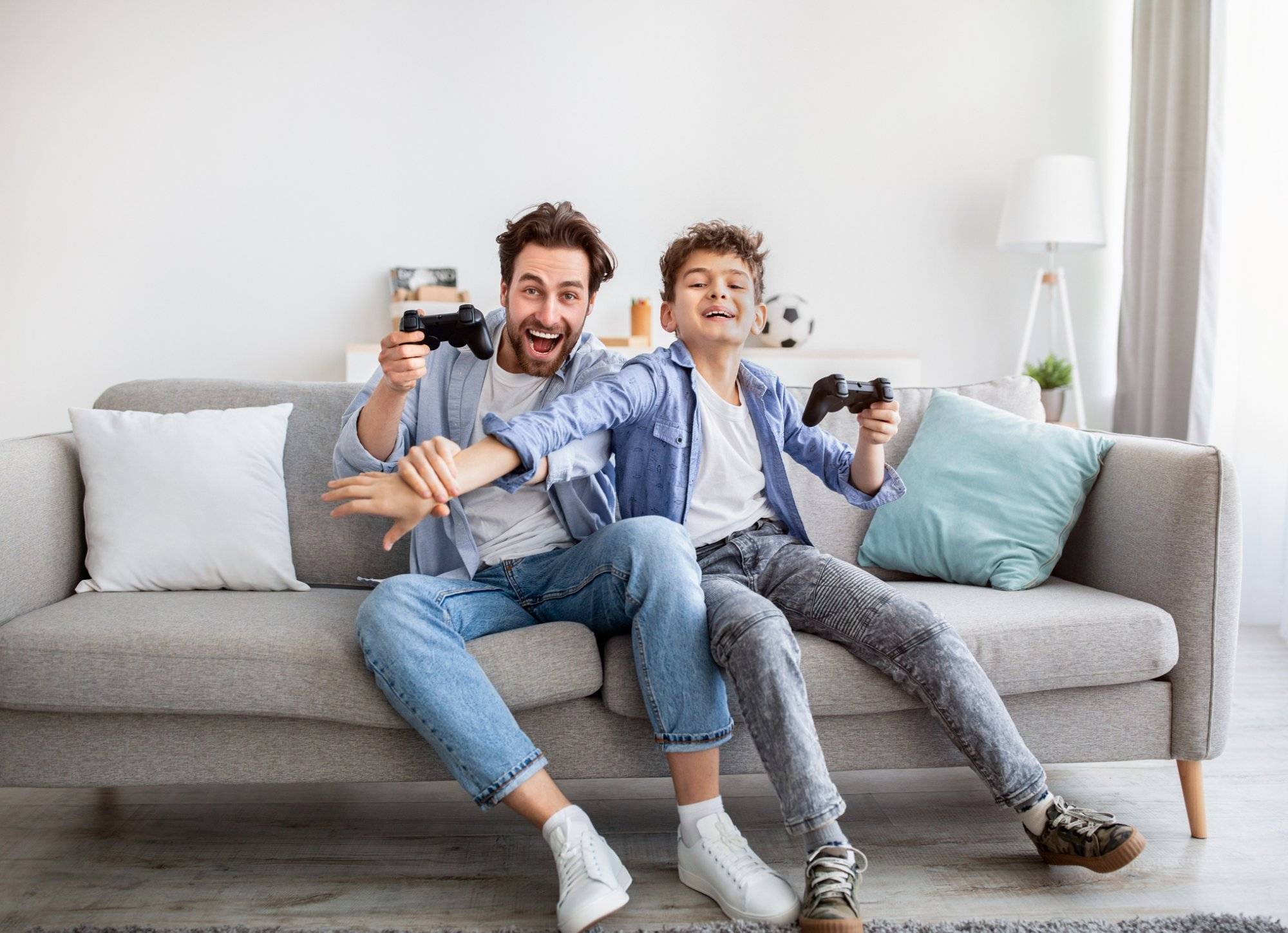 Joyful dad and son with joysticks playing video games at home, boy distracting father with hand