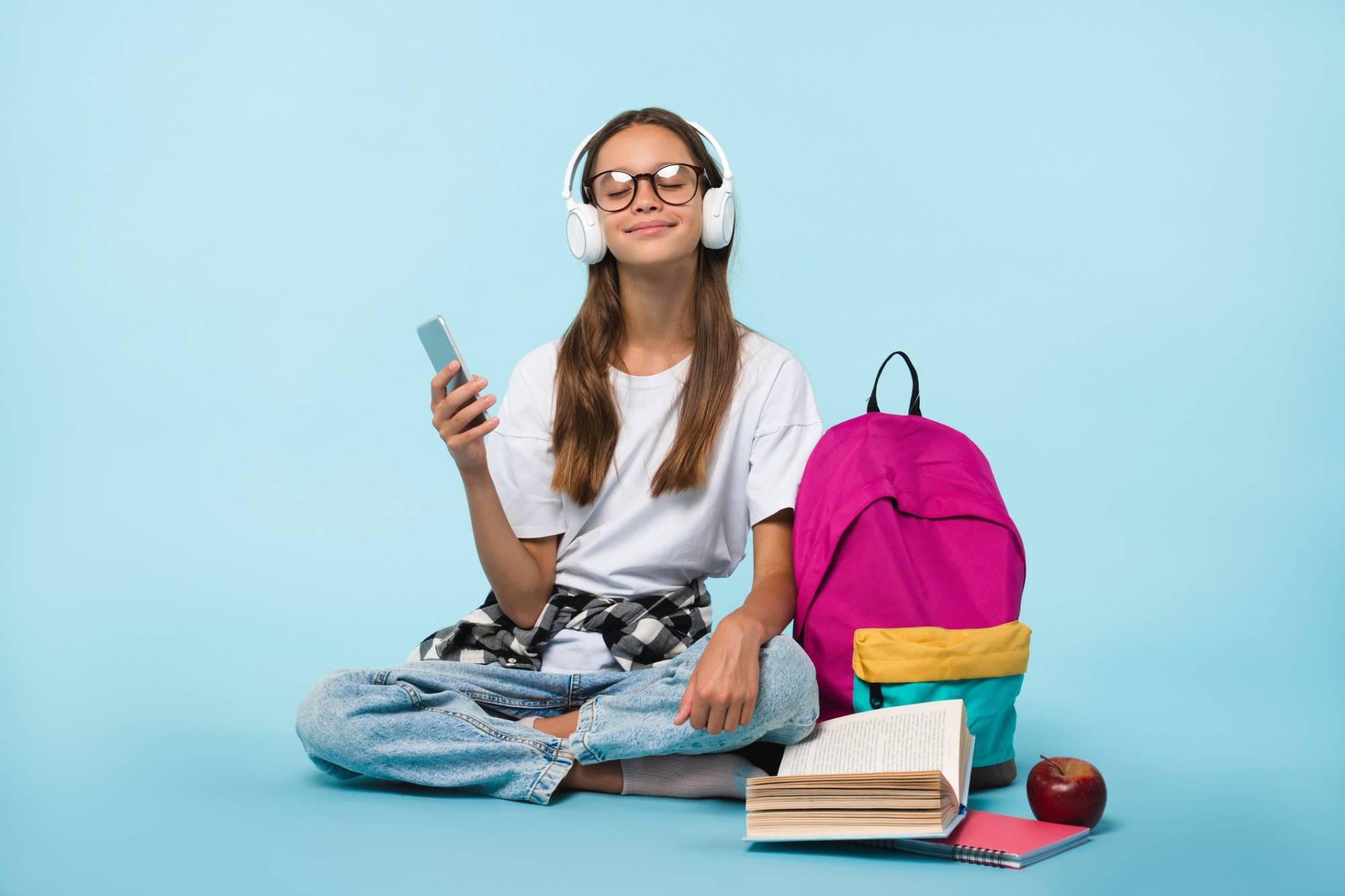 Cheerful caucasian schoolgirl teenager pupil student listening to the music song singer playlist in headphones while preparing going back to school isolated in blue background Cheerful caucasian schoolgirl teenager pupil student listening to the music song singer playlist in headphones while preparing going back to school isolated in blue background