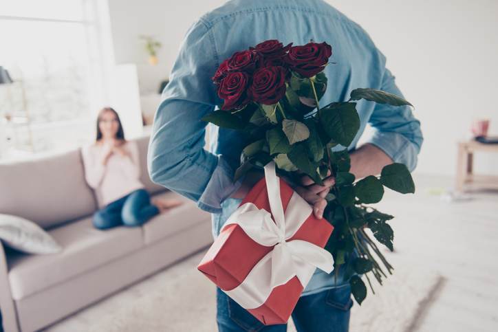 Unexpected moment in routine everyday life! Cropped photo of man's hands hiding holding chic bouquet of red roses and gift with white ribbon behind back, happy woman is on blurred background